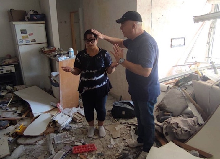 Voorganger Borenstejn uit Ashelon bidt met een gemeentelid in haar appartement dat werd getroffen door een raket uit Gaza. Zelf overleefde de vrouw omdat ze 15 minuten na de aanslag thuis kwam.
